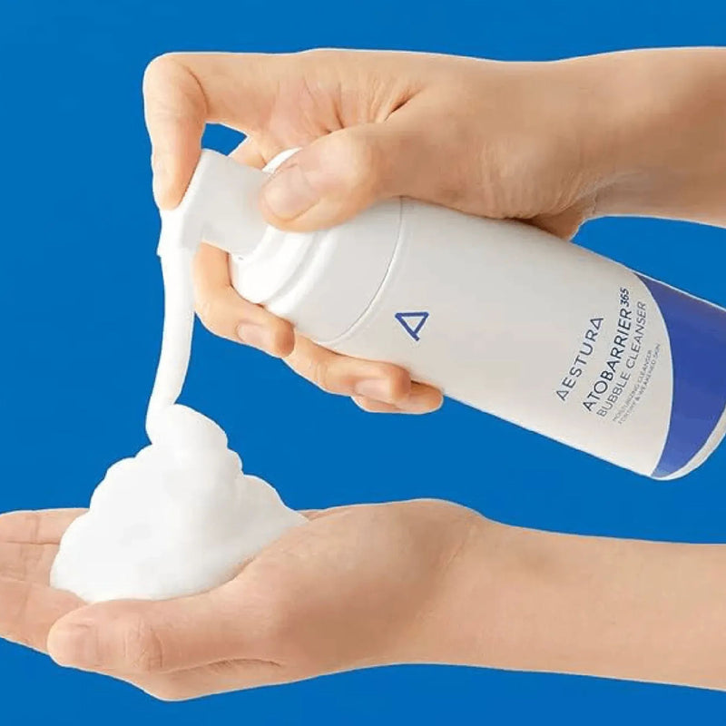 Hand holding a bottle of Aestura AtobARRIER cleanser with foam on a blue background