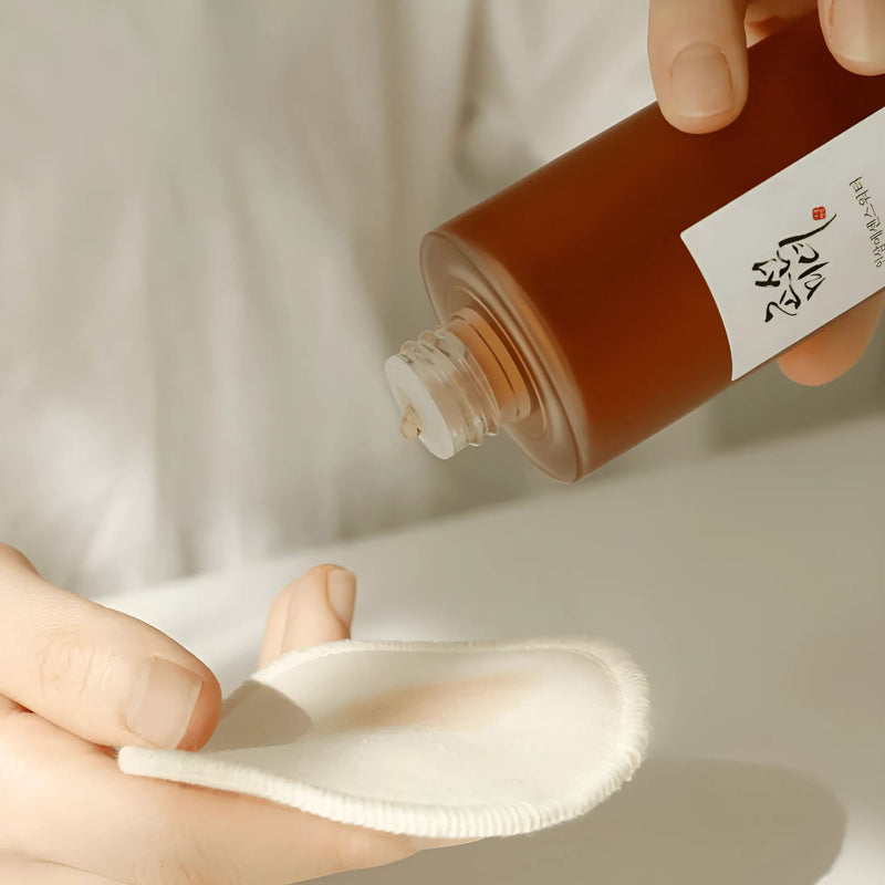 Person applying a brown liquid from a Beauty of Joseon Ginseng Essence Water bottle onto a white cotton pad against a neutral background