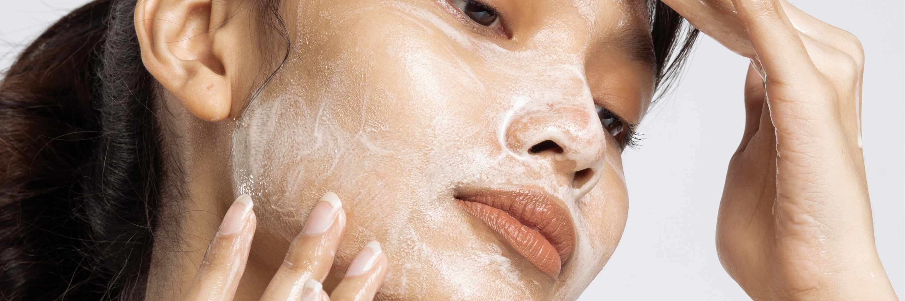 Close-up of a person applying cleanser to their face with a neutral background