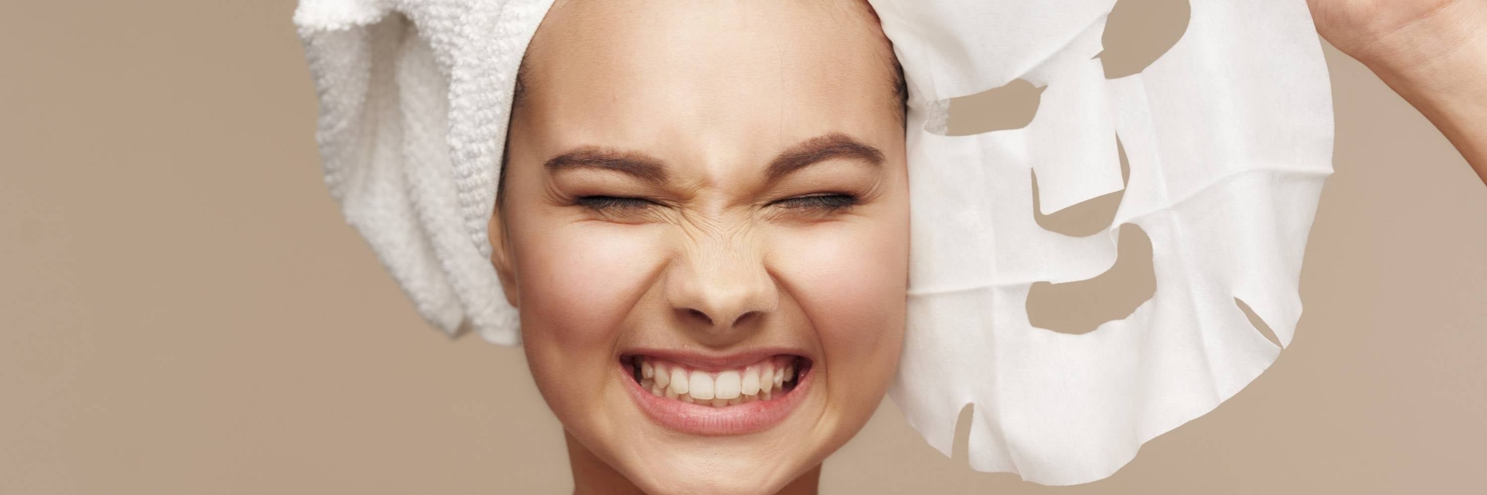 Woman applying a facial mask with a towel wrapped around her head against a beige background.