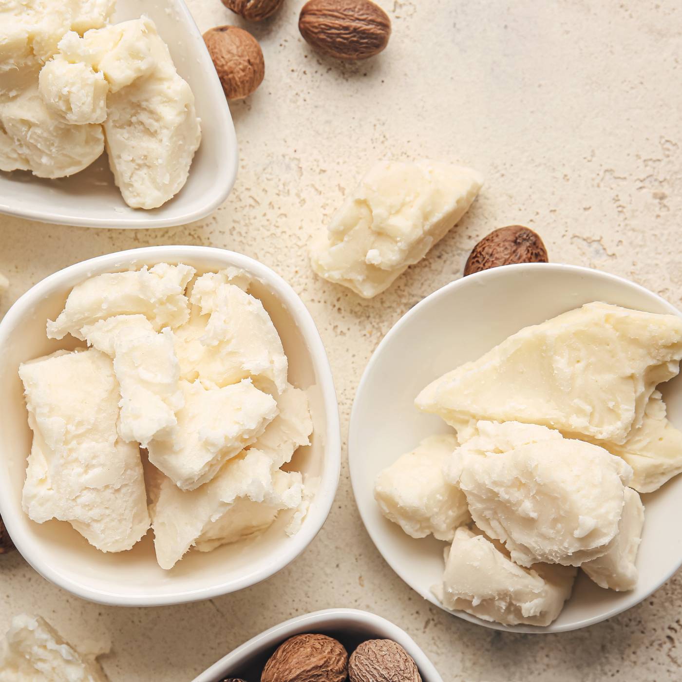 Three white bowls filled with shea butter on a light brown surface.