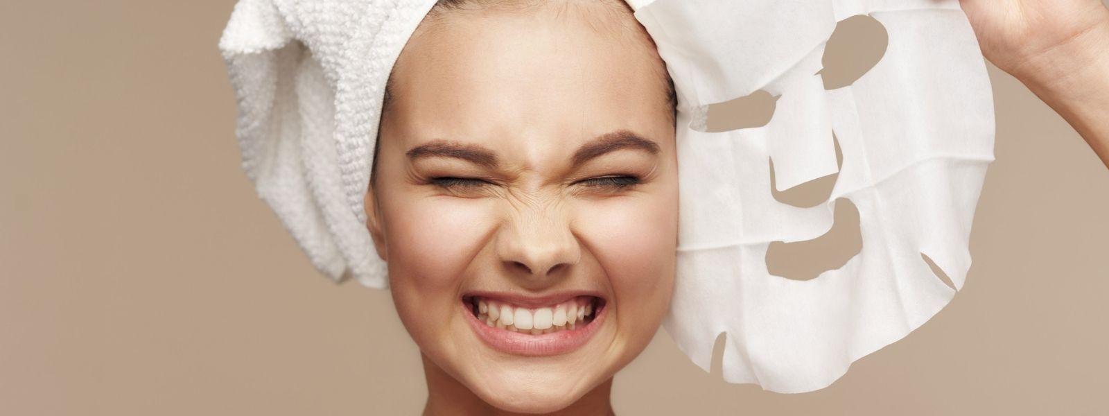 Woman with a towel on her head holding a sheet mask against a beige background