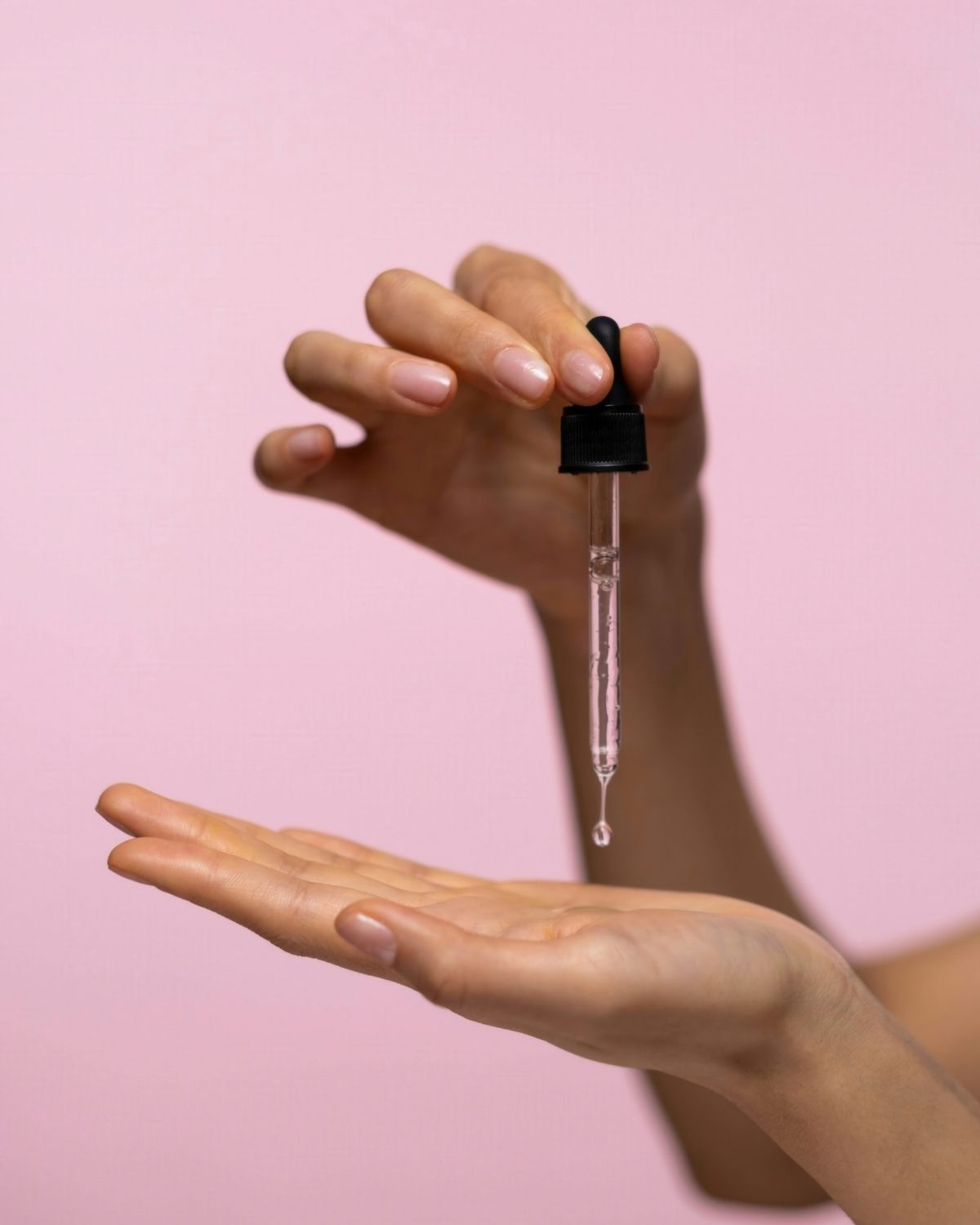 Hand holding a dropper with a clear liquid over another hand on a pink background