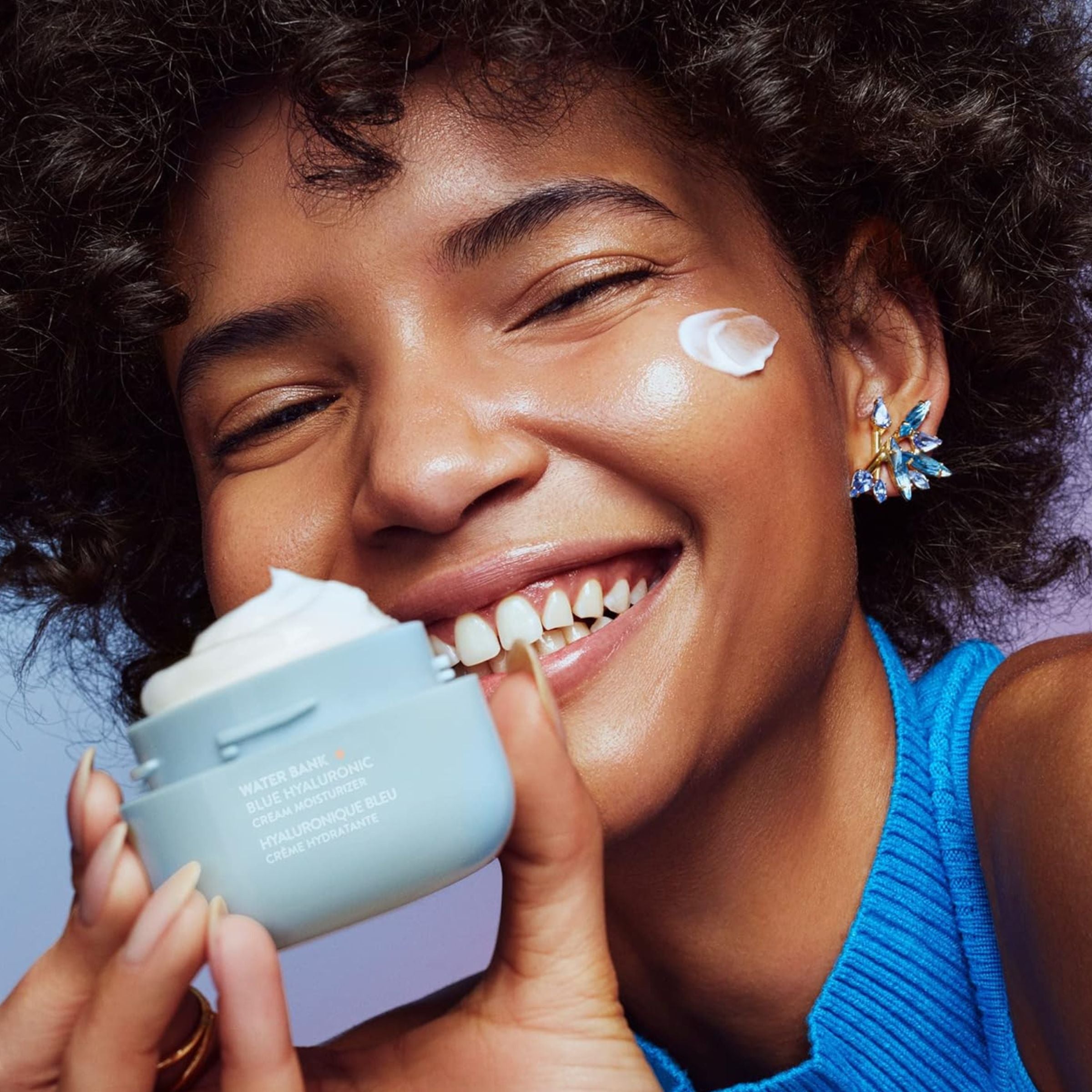 Woman applying cream to her face with a jar of cream in her hand, against a light purple background.