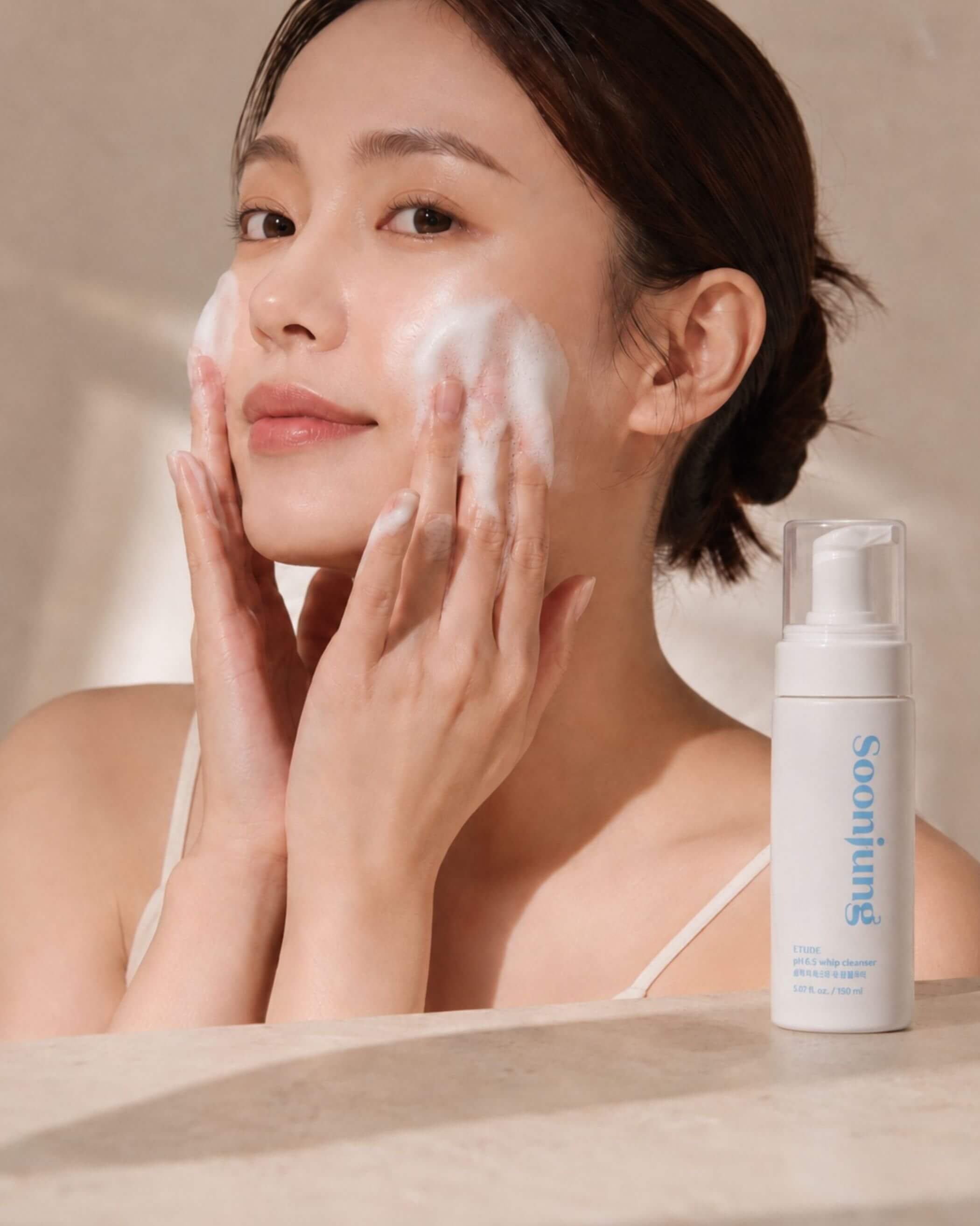 Woman applying cleanser to her face with a bottle of Etude SoonJung pH 6.5 Whip Cleanser in the foreground.