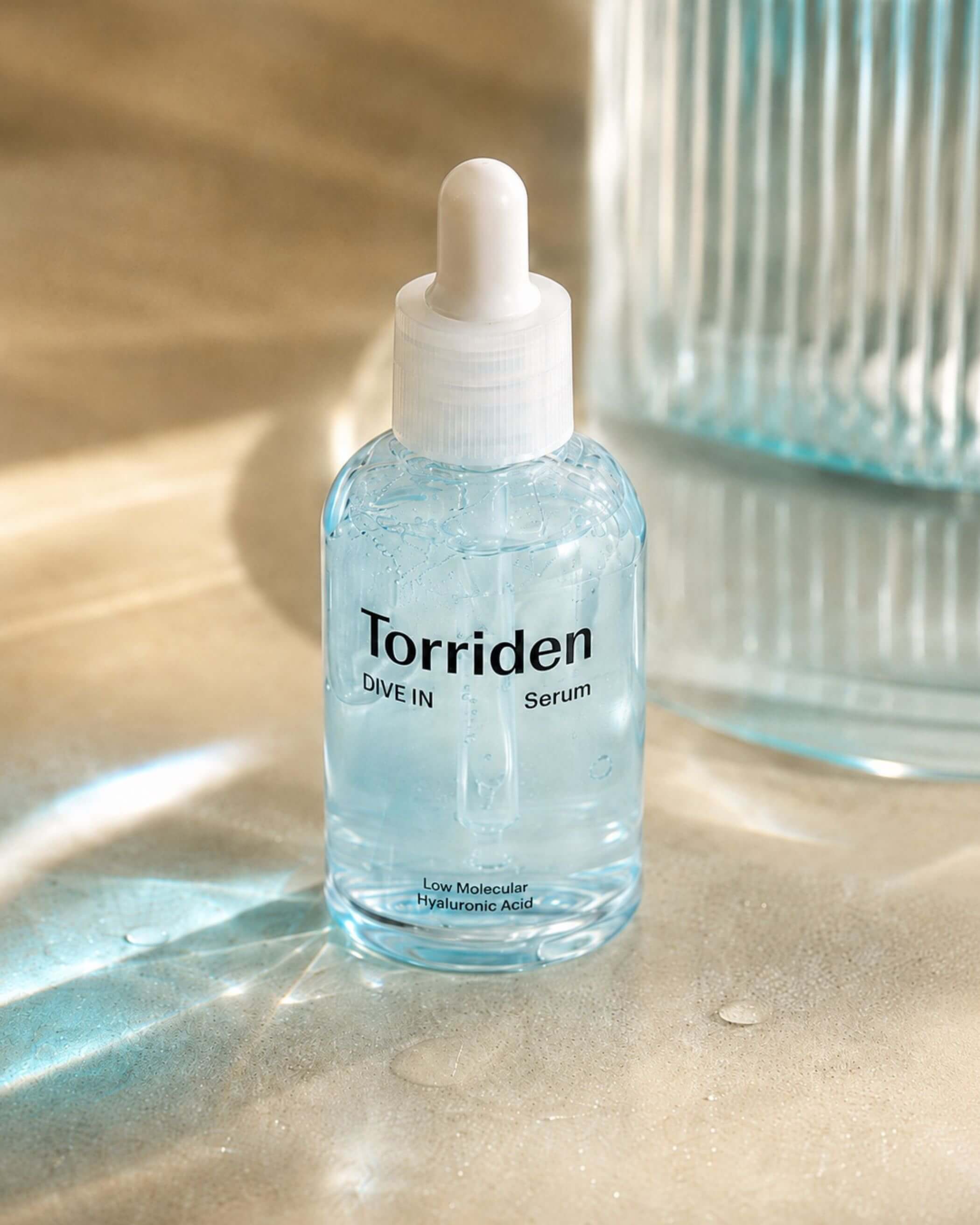 Torriden Dive In Serum bottle on a textured surface with a blurred glass container in the background