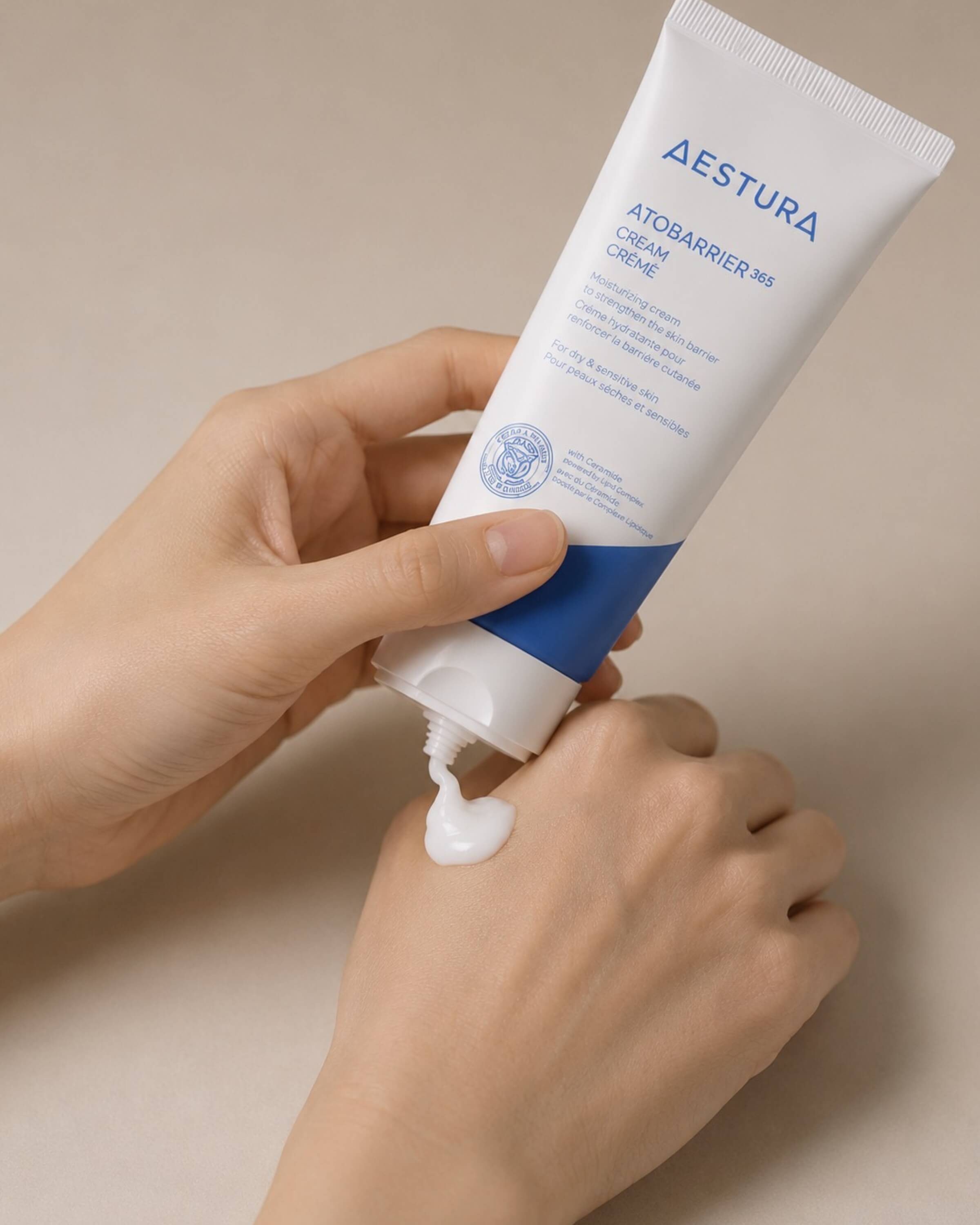 Hand holding a tube of Aestura Atobarrier 365 cream against a beige background