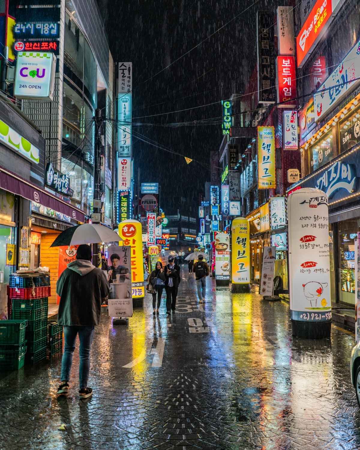 Korean street at night with illuminated signs and people with umbrellas.